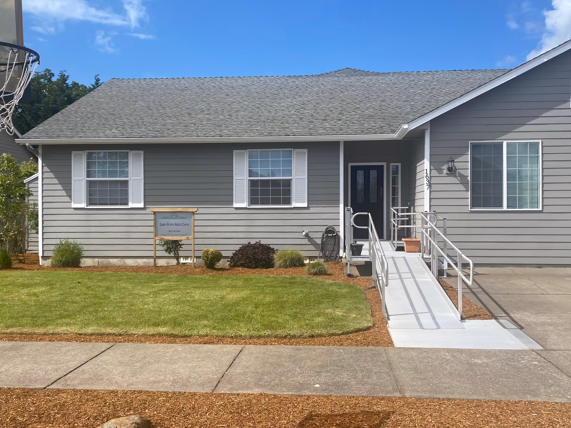 Front exterior of a single-story gray house with a wheelchair ramp, small lawn, and a sign that reads 'Open Arms Adult Care'.