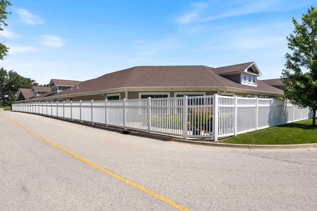 Single-story memory care building with a long white fence along a paved road under a blue sky.