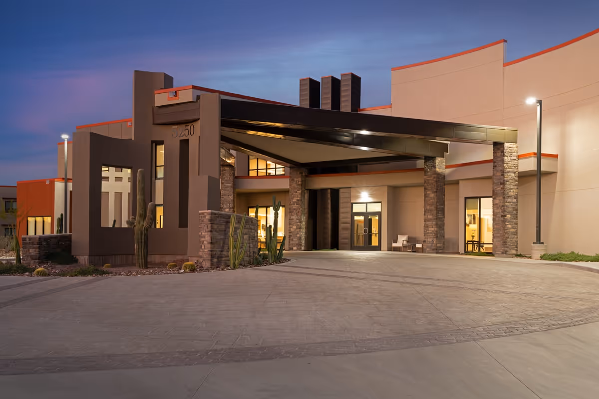 The covered entrance and porte-cochere of a modern senior living building with stone columns and desert landscaping at dusk.