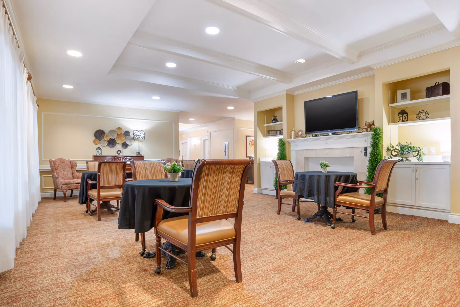 A well-lit common area with several tables covered in black tablecloths, each surrounded by wooden chairs with striped upholstery. The room features a beige carpet, cream-colored walls, a flat-screen TV mounted above a white fireplace, built-in shelves with decorative items, and a large window with sheer white curtains allowing natural light to enter.