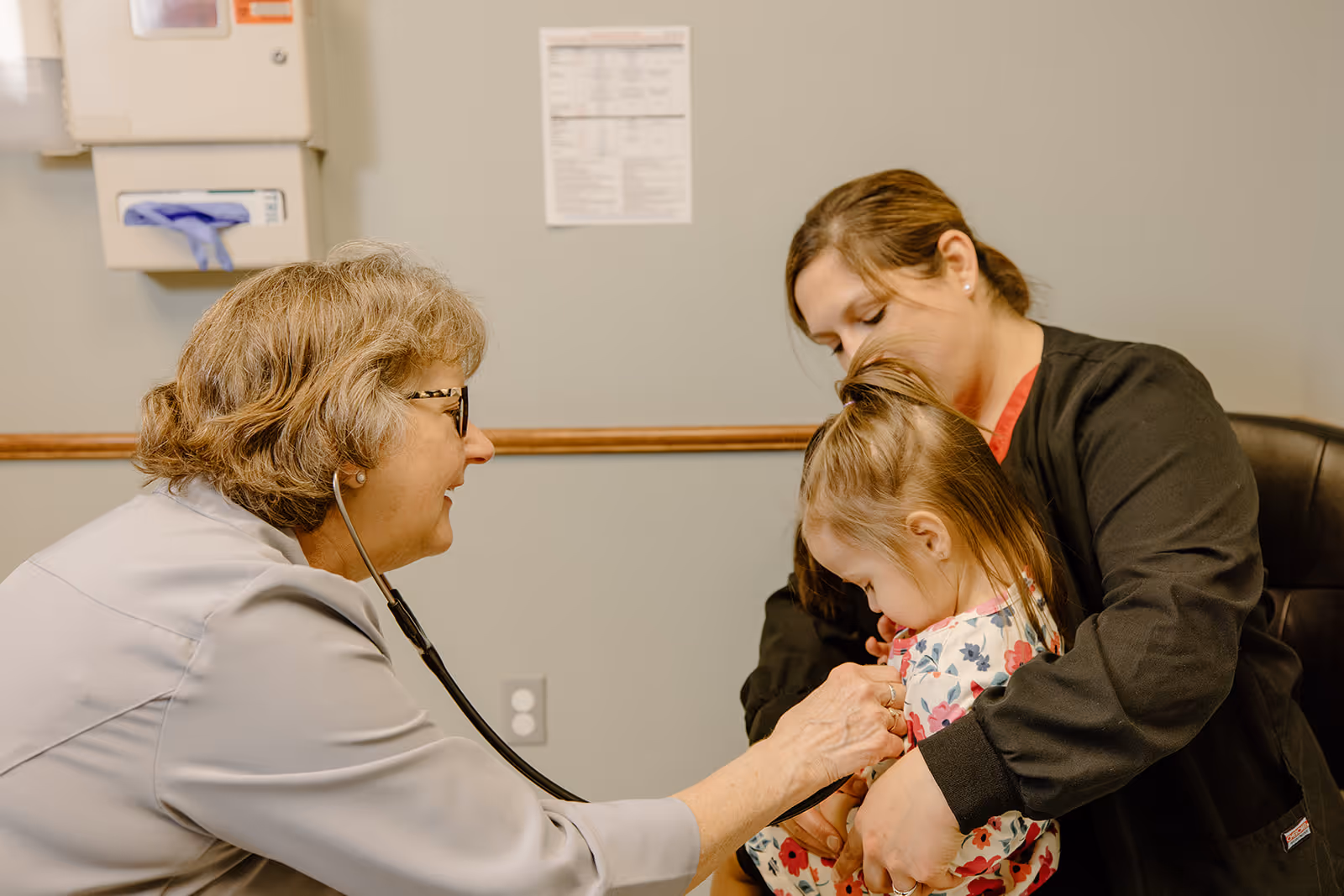 A healthcare professional uses a stethoscope to listen to the chest of a young girl who is sitting on a woman's lap in a medical examination room.