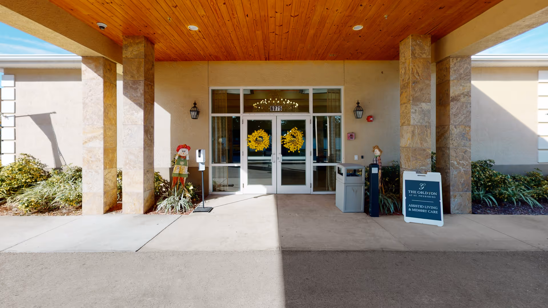 Entrance of The Goldton at St. Petersburg facility with double glass doors decorated with yellow wreaths. The entrance is covered by a wooden ceiling supported by stone pillars. There are two scarecrow decorations on either side of the doors, a hand sanitizer dispenser, a trash bin, and a signboard displaying the facility name and services.