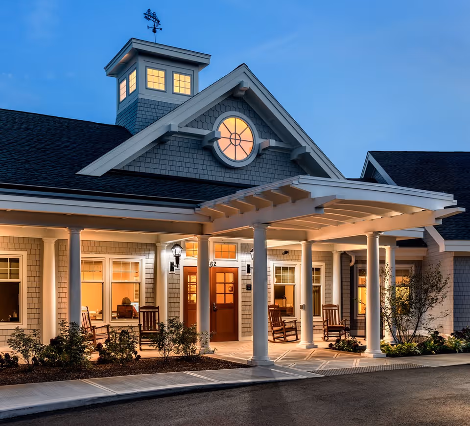 Exterior view of a senior living facility entrance at dusk with warm lights glowing from inside. The building features a covered porch supported by white columns, rocking chairs on the porch, and a decorative cupola with windows and a weather vane on the roof.