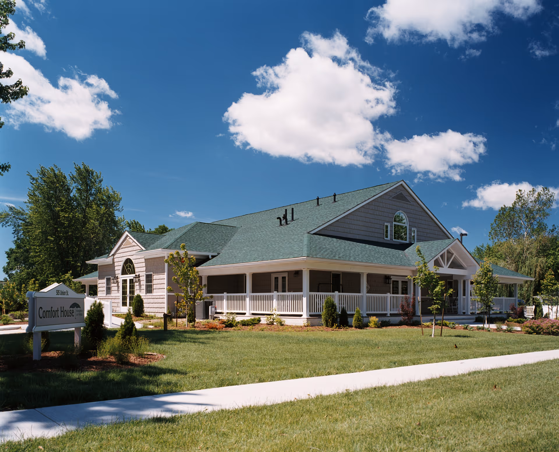 Exterior view of a single-story senior living facility with a wraparound porch and green lawn.