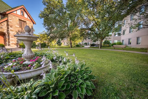 Sunny courtyard with a stone fountain planter surrounded by flowering plants, lawns, trees, benches, and adjacent multi-story buildings.