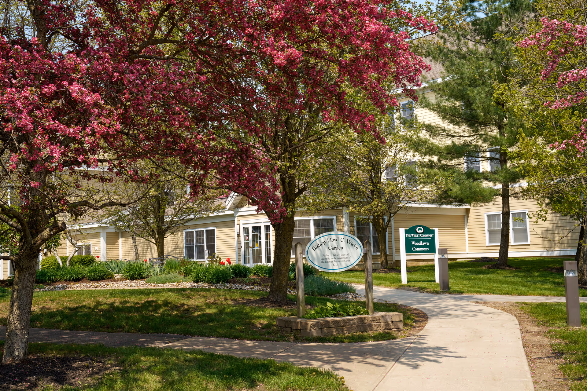 Exterior front of a beige senior living building with a curved walkway, flowering trees, and signs for The Wesley Community - Woodlawn Commons.