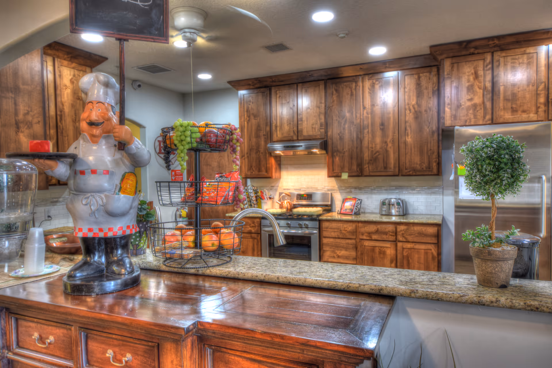A kitchen area with wooden cabinets, a stainless steel refrigerator, a stove with a range hood, and a granite countertop. On the counter, there is a decorative chef statue holding a tray, a tiered fruit basket with grapes and apples, a small potted plant, and a water dispenser with cups.