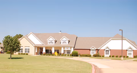 Exterior view of a single-story senior living facility building with a combination of brick and light-colored siding, multiple windows, a covered entrance, and a well-maintained lawn with a tree and a curved driveway under a clear blue sky.