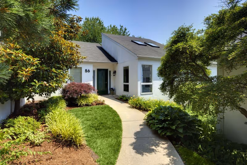 Front entrance of a white single-story cottage numbered 56 with a curved concrete walkway and lush landscaped plants.