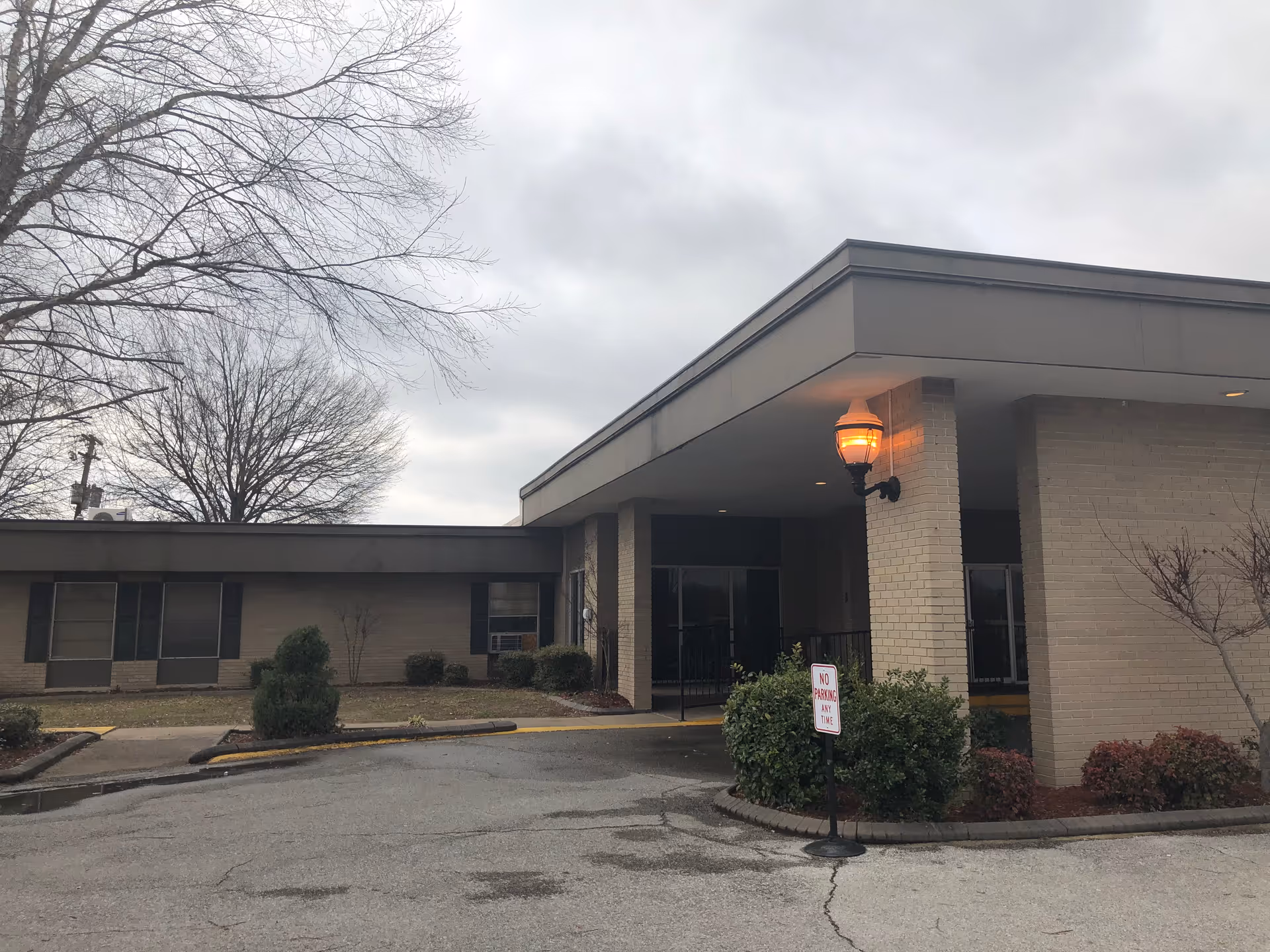 Front entrance with a covered canopy, lit lantern, and brick facade of the senior living facility under an overcast sky.