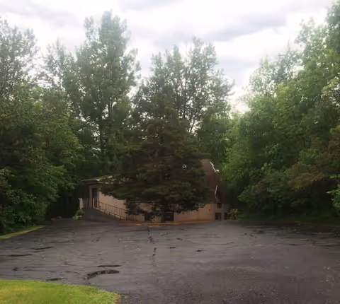 Front of a small building partially hidden by trees, with a large paved parking lot.
