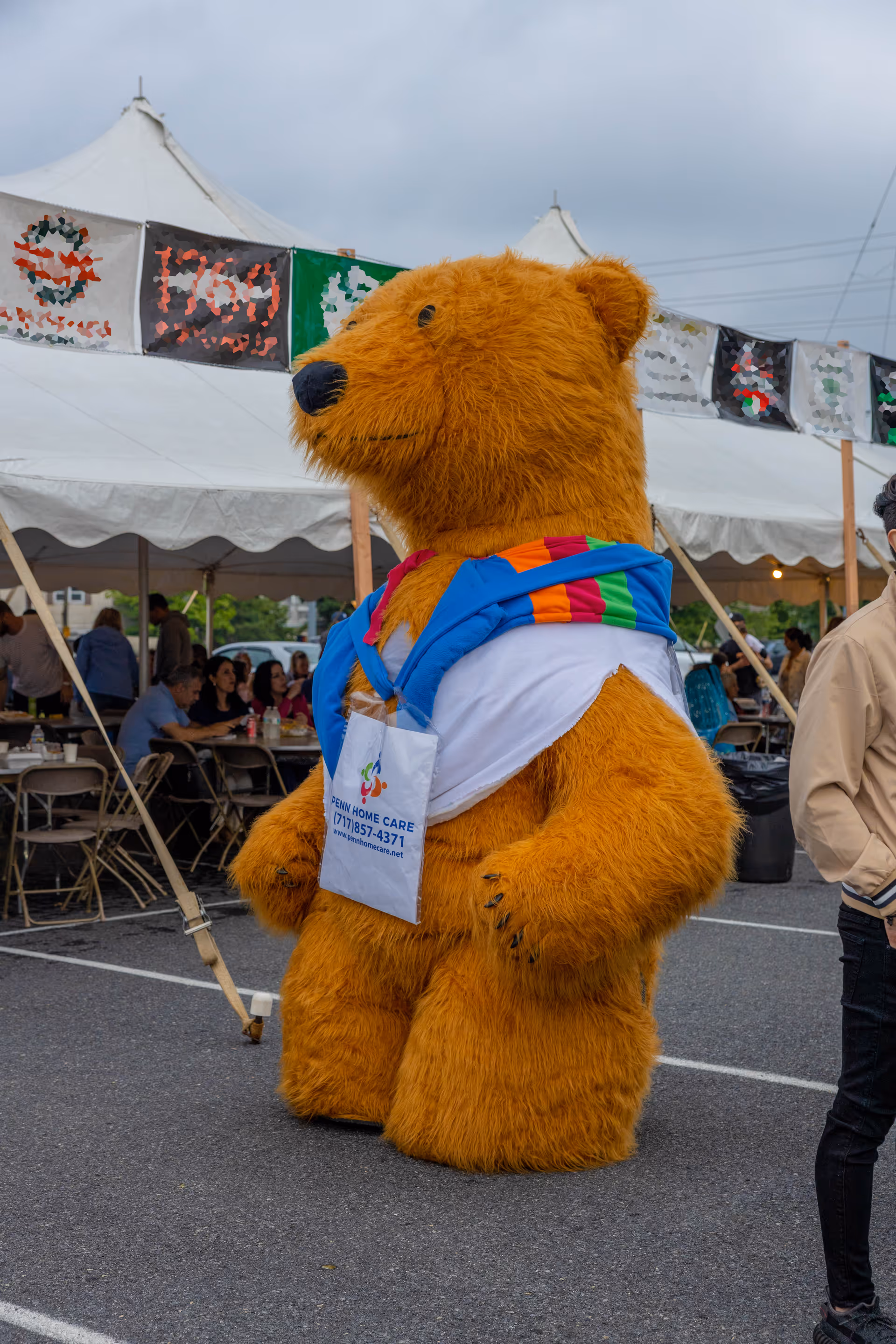 An orange bear mascot wearing a colorful scarf stands outdoors at a Penn Home Care event.