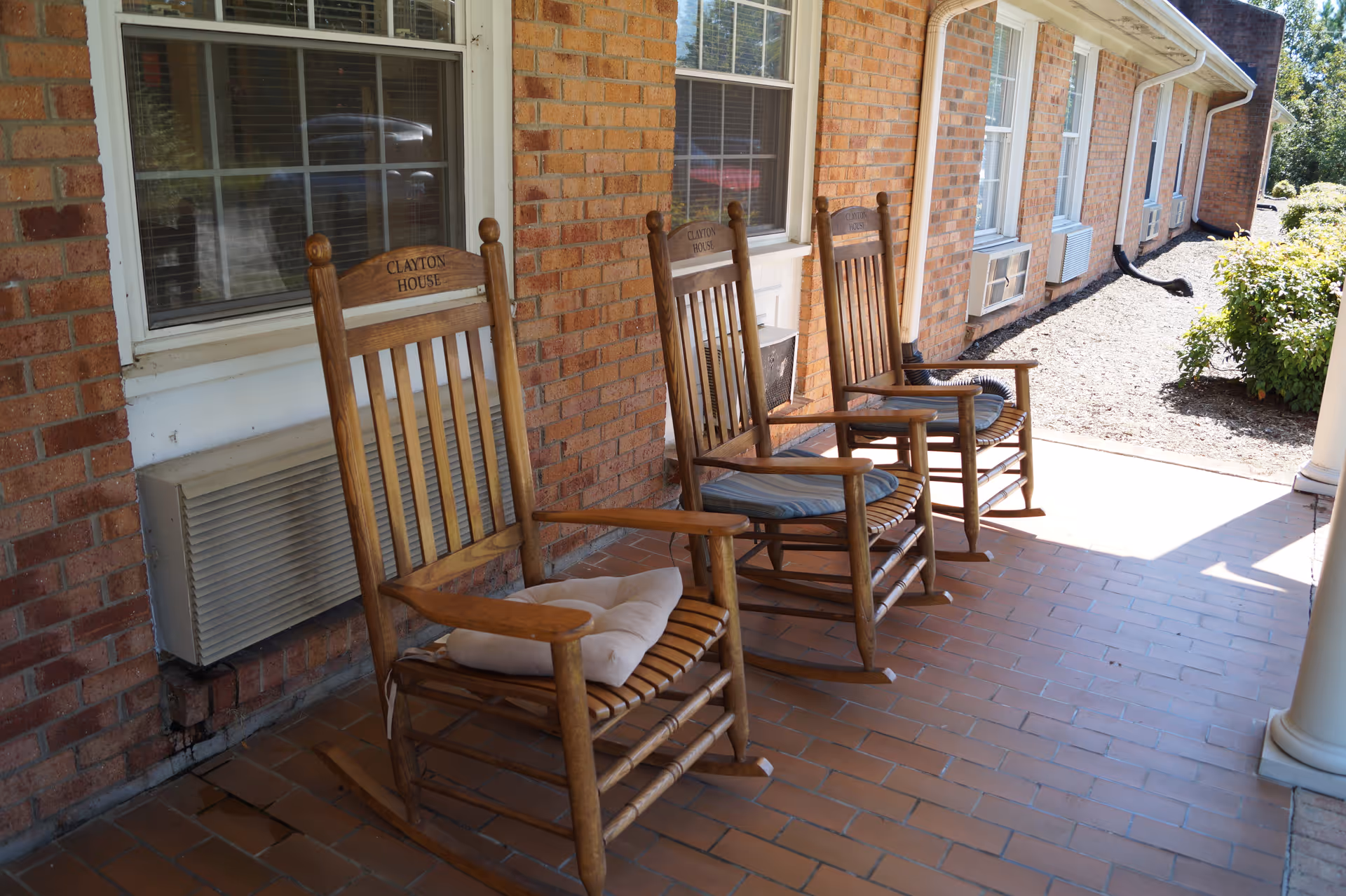 Three wooden rocking chairs with cushions on a brick front porch of the Clayton House.
