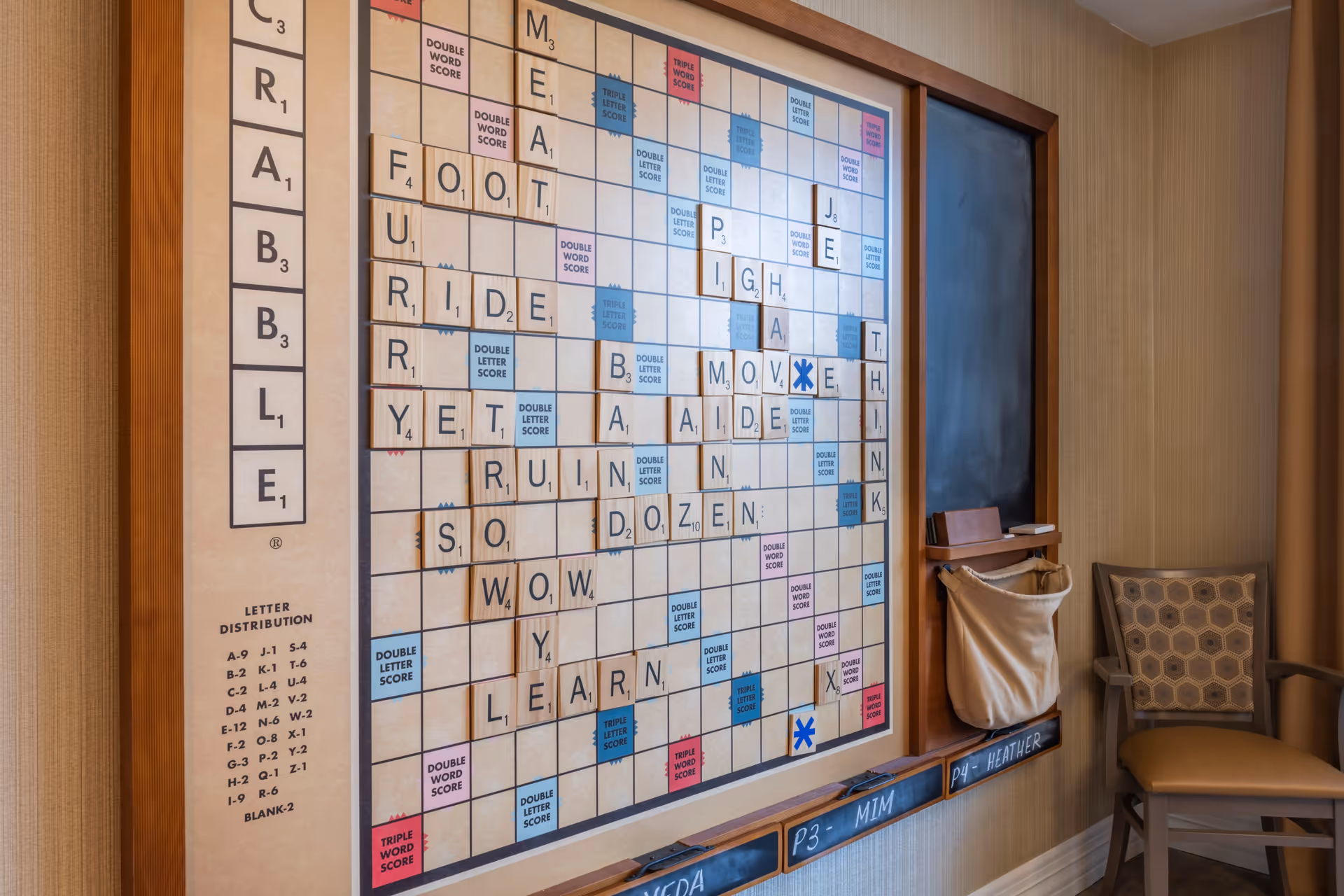 A large wall-mounted Scrabble game board with wooden letter tiles forming various words. To the right of the board is a chalkboard with a small basket attached below it, and a patterned cushioned chair is positioned nearby. The setting appears to be a cozy indoor recreational area.