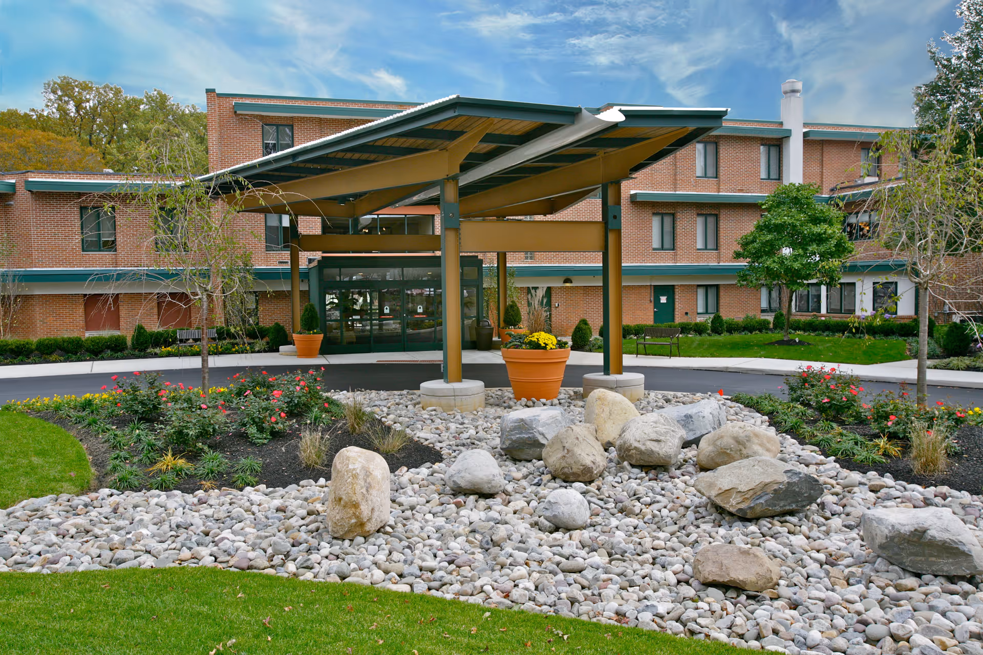 Front entrance of a senior living facility with a covered drop-off area, landscaped garden with rocks and flowers, and a three-story brick building in the background under a partly cloudy sky.