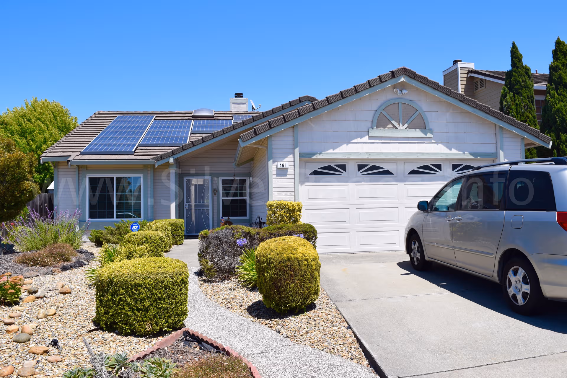Sunlit front exterior of a suburban house with solar panels and a silver minivan in driveway.