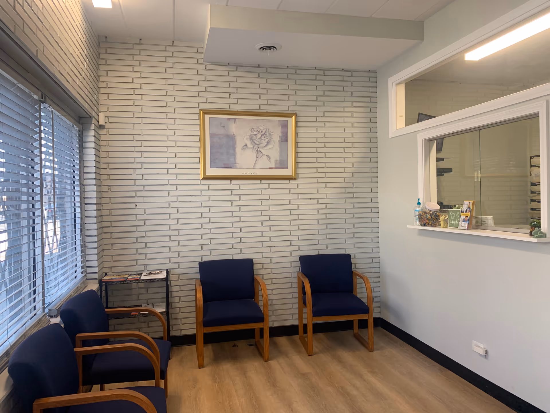 Quiet waiting area with blue chairs, a white brick wall, and a reception window.