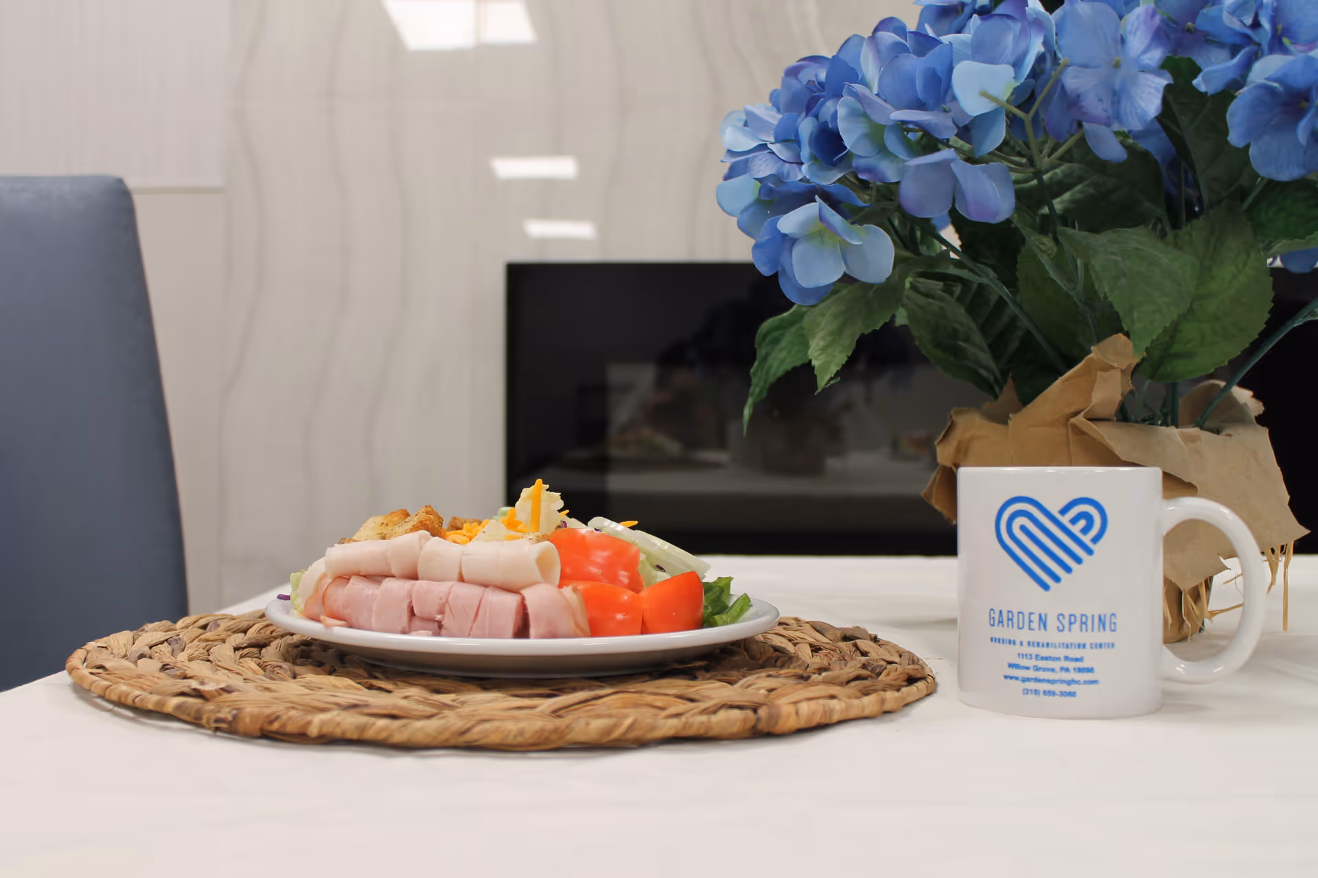 A dining table with a plate of sliced ham and vegetables, blue flowers, and a mug.