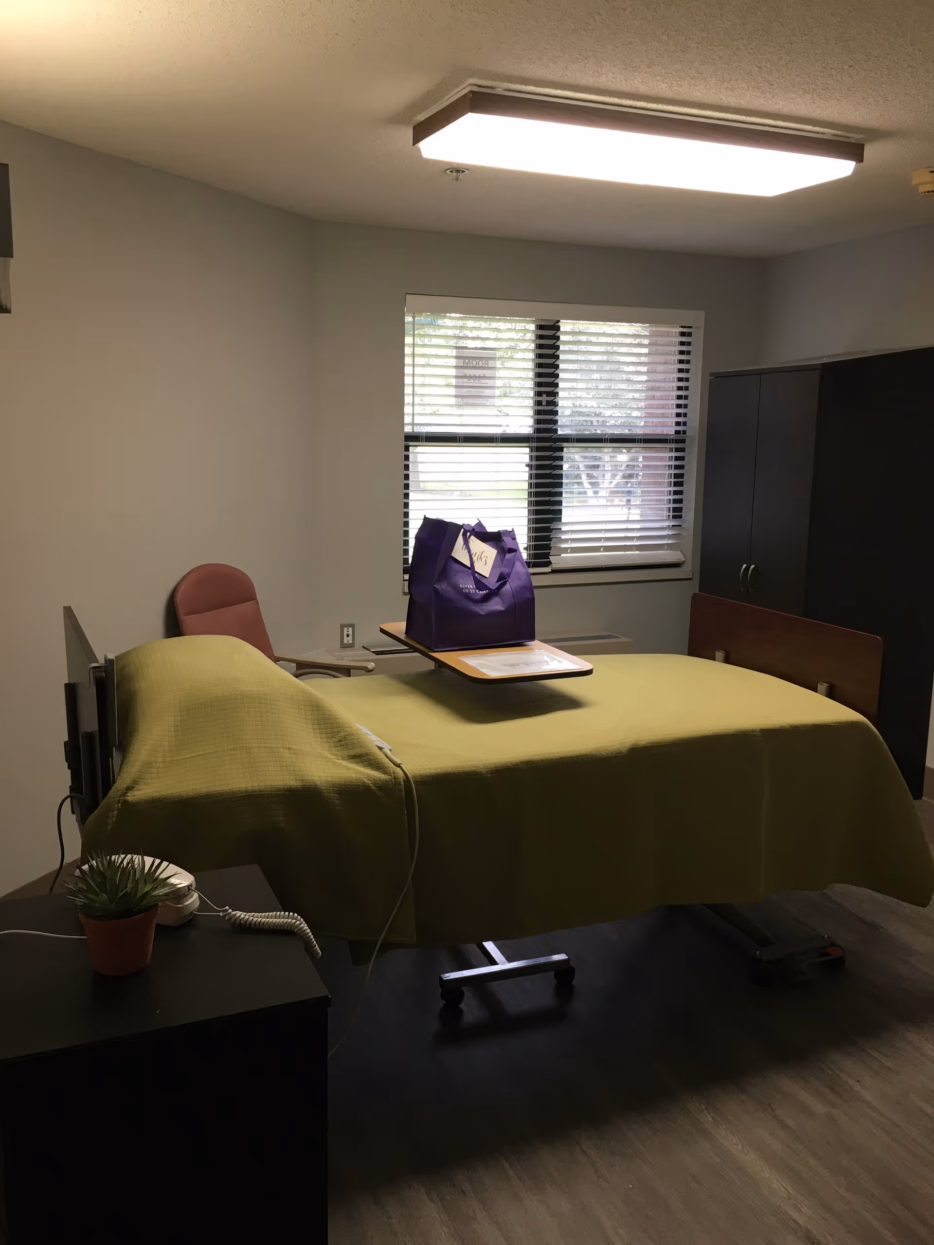 Senior living bedroom with a bed, green cover, window with blinds, and a purple bag.