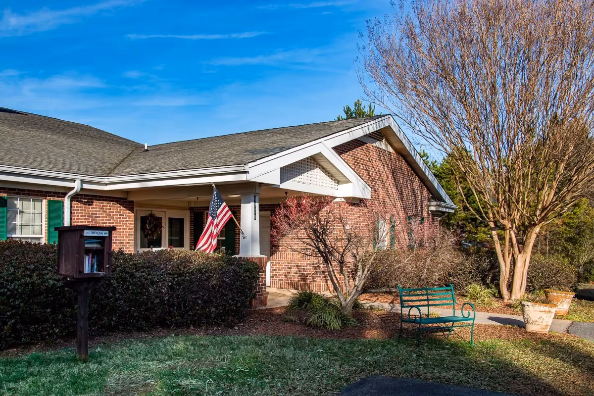 Brick senior living building entrance with American flag, shrubs, and a green bench outside.
