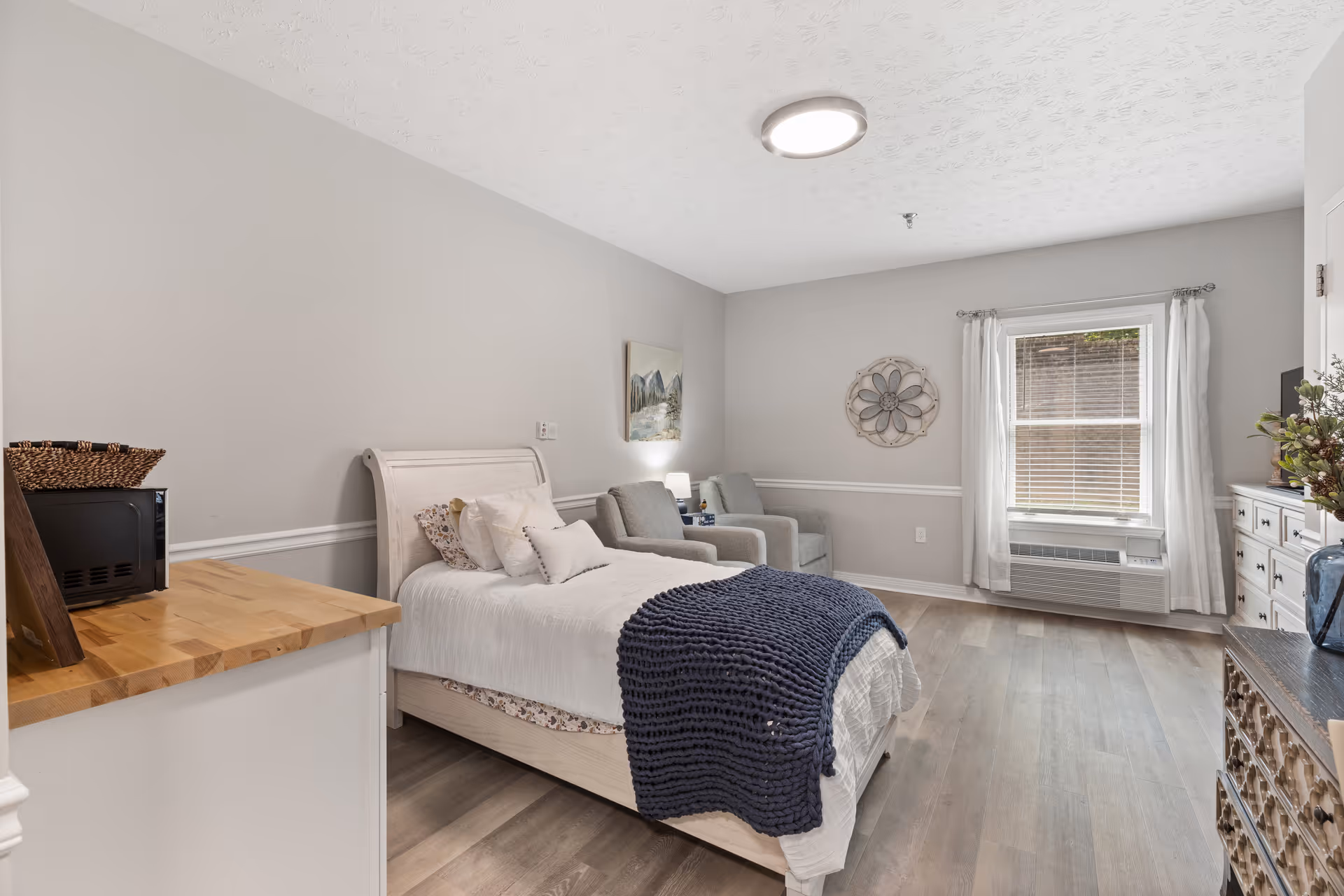 Bright, modern bedroom with a bed, seating area, dresser, and a large window.