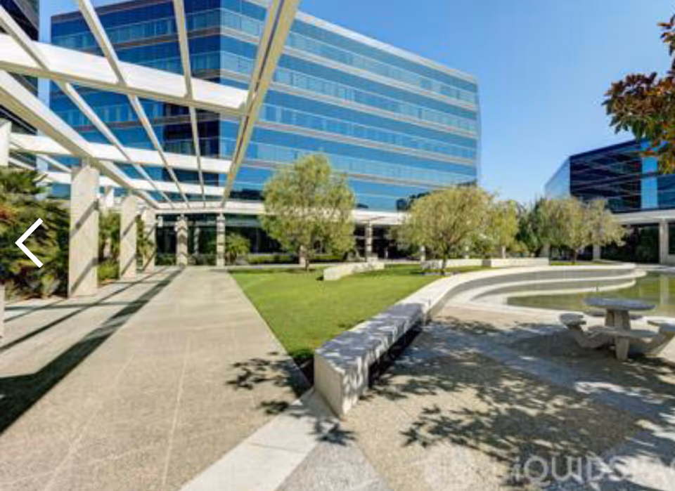 Modern glass buildings surround a sunny outdoor courtyard with trees and seating.