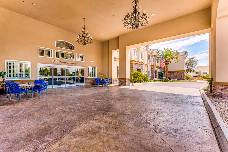 Covered entrance area of a senior living facility with a high ceiling, chandeliers, and seating arrangements with blue chairs and tables. The entrance doors have a 'Welcome Home' sign above them. Outside, there are palm trees, an American flag, and part of the building exterior visible under a clear sky.