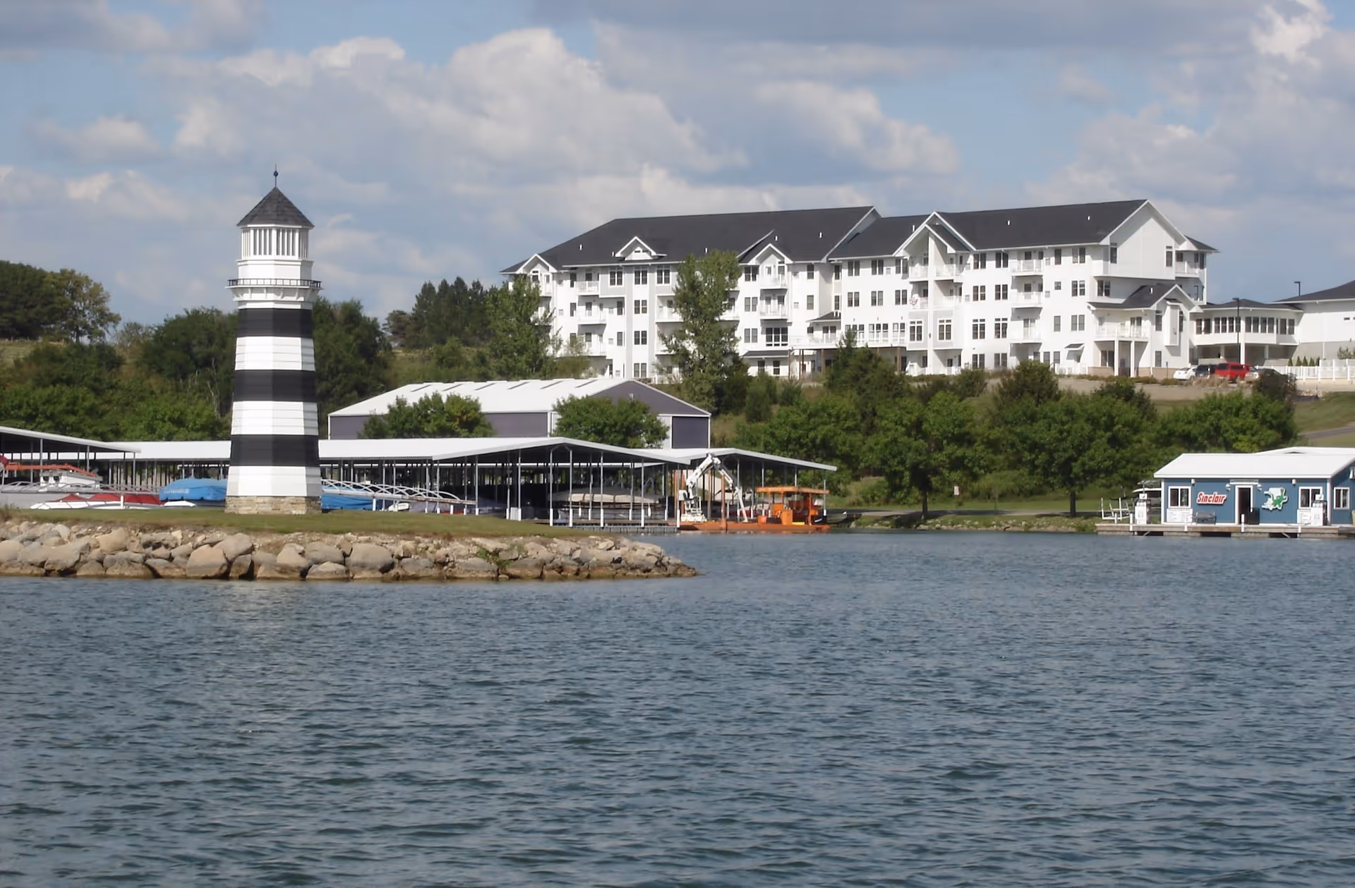 View of a waterfront senior living facility named The Lakeside Village, featuring a large white multi-story building with balconies, a black and white striped lighthouse on a rocky shore, covered boat docks, and a small blue building near the water under a partly cloudy sky.