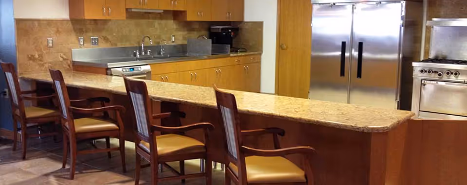A communal kitchen with a long granite counter and several wooden bar chairs in front of stainless steel appliances and a sink.