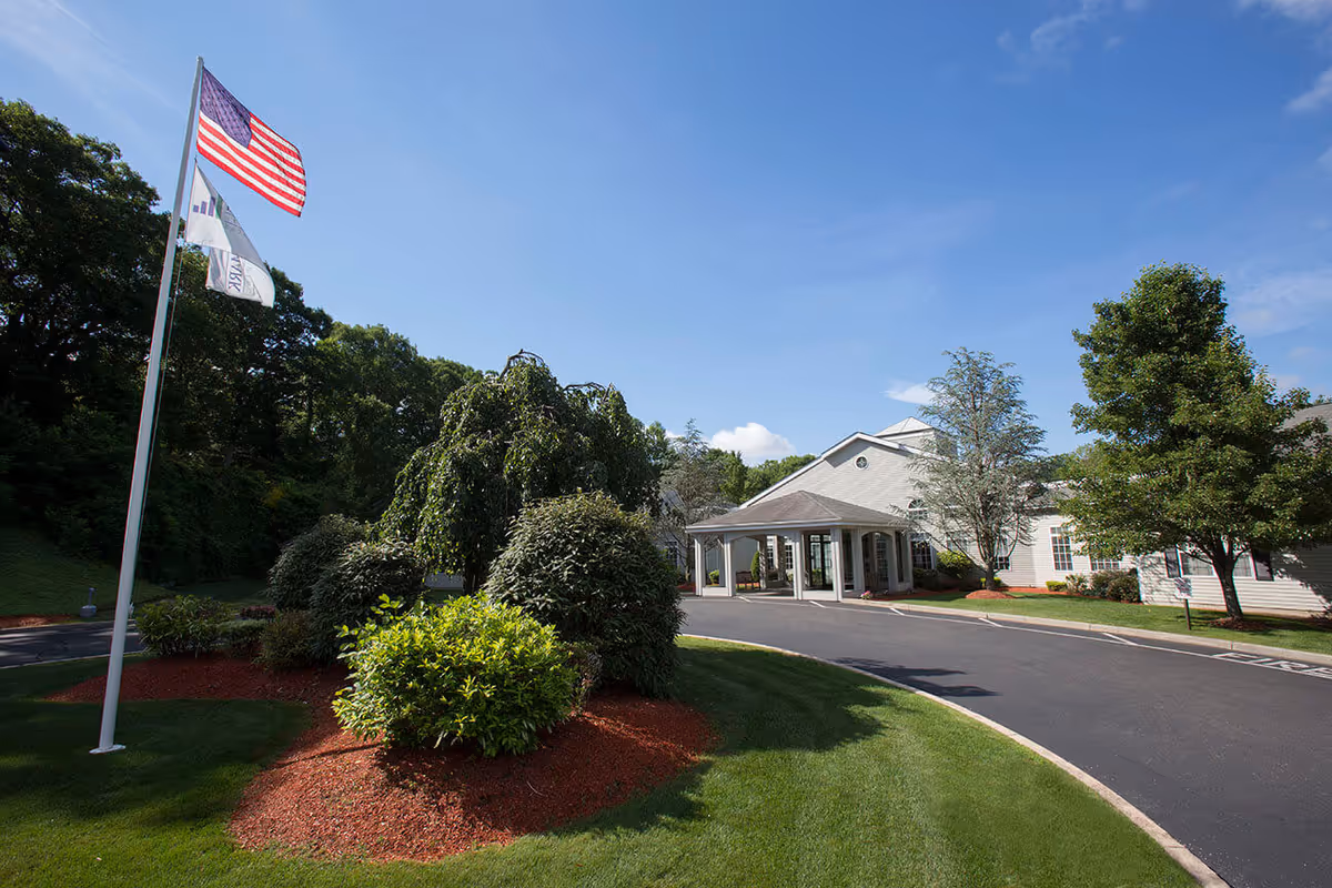 Front entrance of a senior living building with a covered drop-off, driveway, landscaped shrubs, and flagpoles under a blue sky.