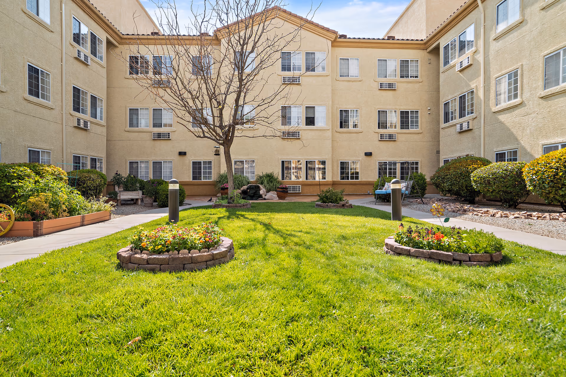 A sunny courtyard area with green grass, two circular flower beds with colorful flowers, a leafless tree in the center, and beige multi-story building walls with many windows surrounding the courtyard. There are bushes and plants along the edges and a paved walkway.