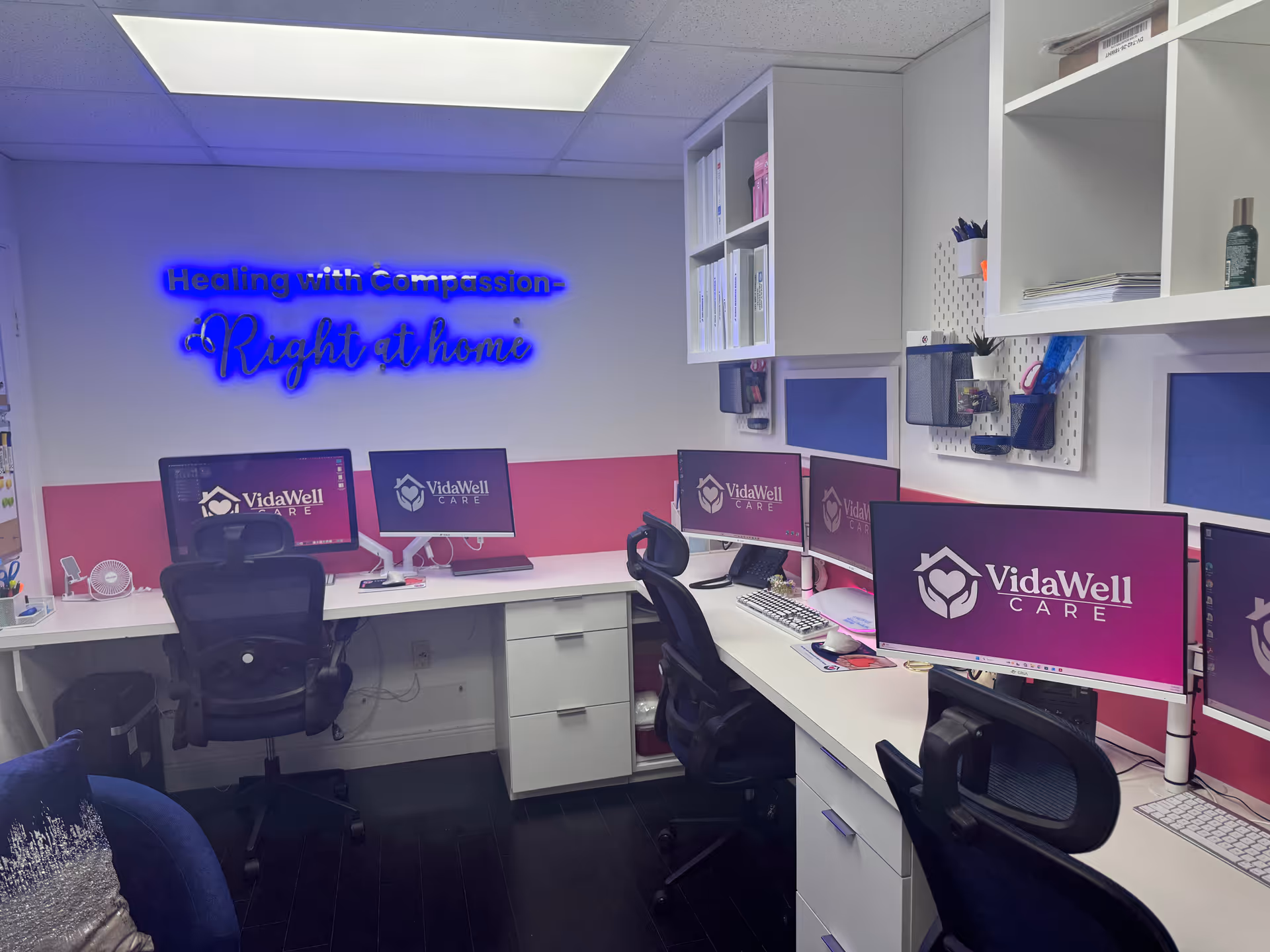 A bright office with multiple VidaWell monitors and a neon sign on the wall.