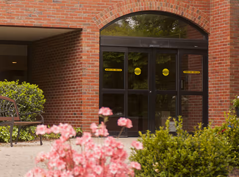 Brick building entrance with glass doors, yellow signs, and blooming pink flowers.