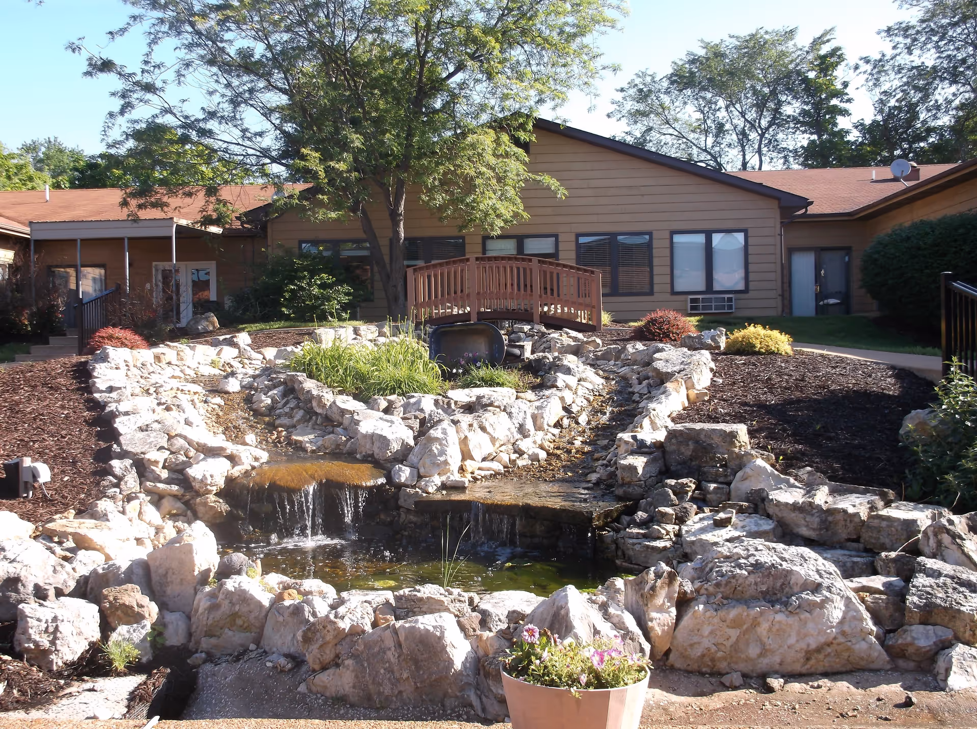 Outdoor garden area at StoneBridge Senior Living - De Soto featuring a small waterfall flowing over rocks into a pond, surrounded by landscaping with plants and mulch. A wooden footbridge crosses over the rocky water feature, with a building and trees in the background under a clear sky.