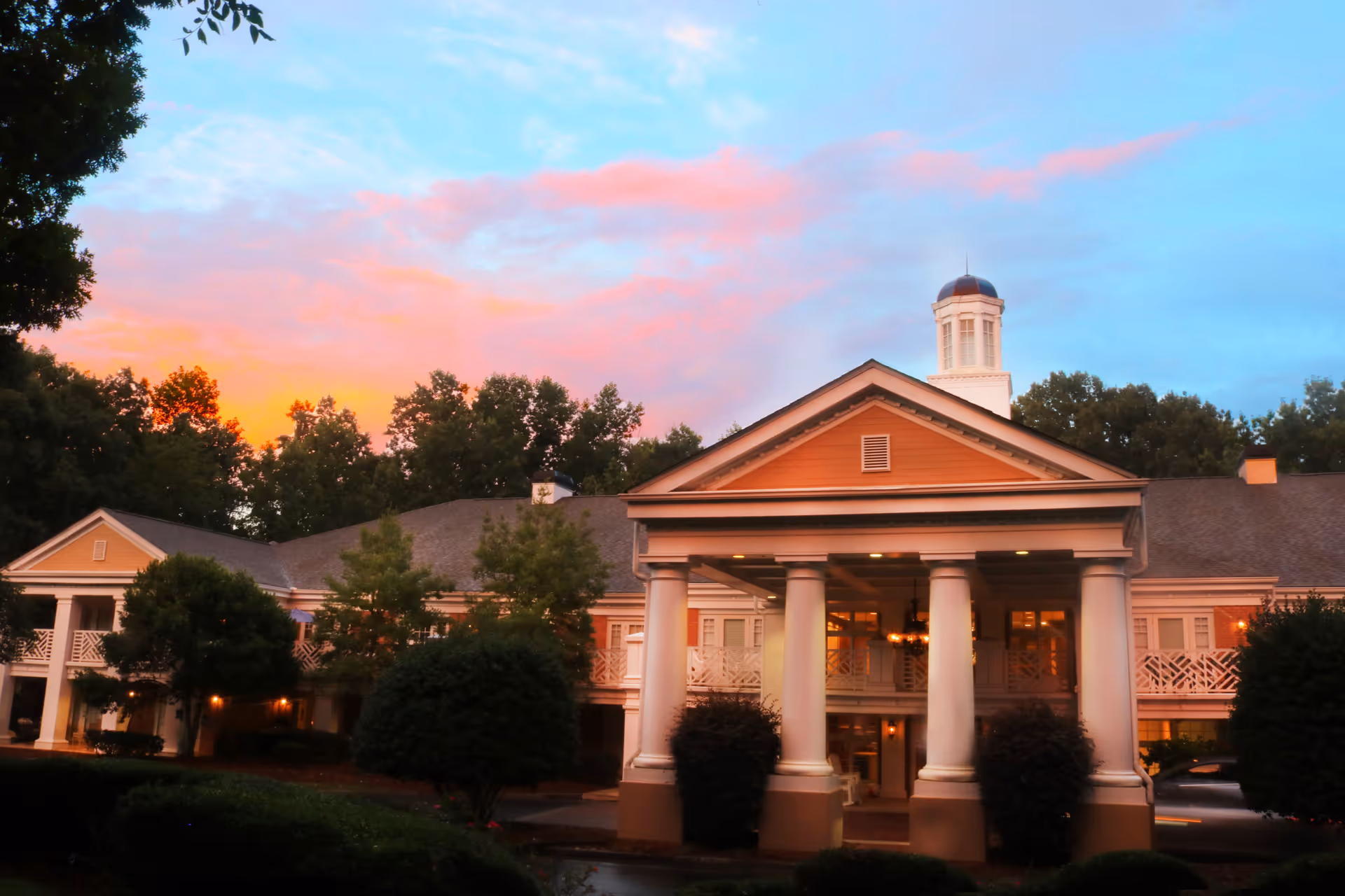 Exterior view of a large assisted living facility building with classical architecture, featuring tall white columns at the entrance, a cupola on the roof, surrounded by trees and bushes under a colorful sunset sky.