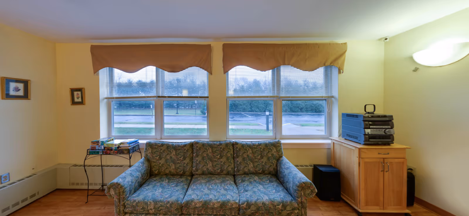 Light-yellow living room with a floral sofa, windows, and a stereo on a wooden cabinet.