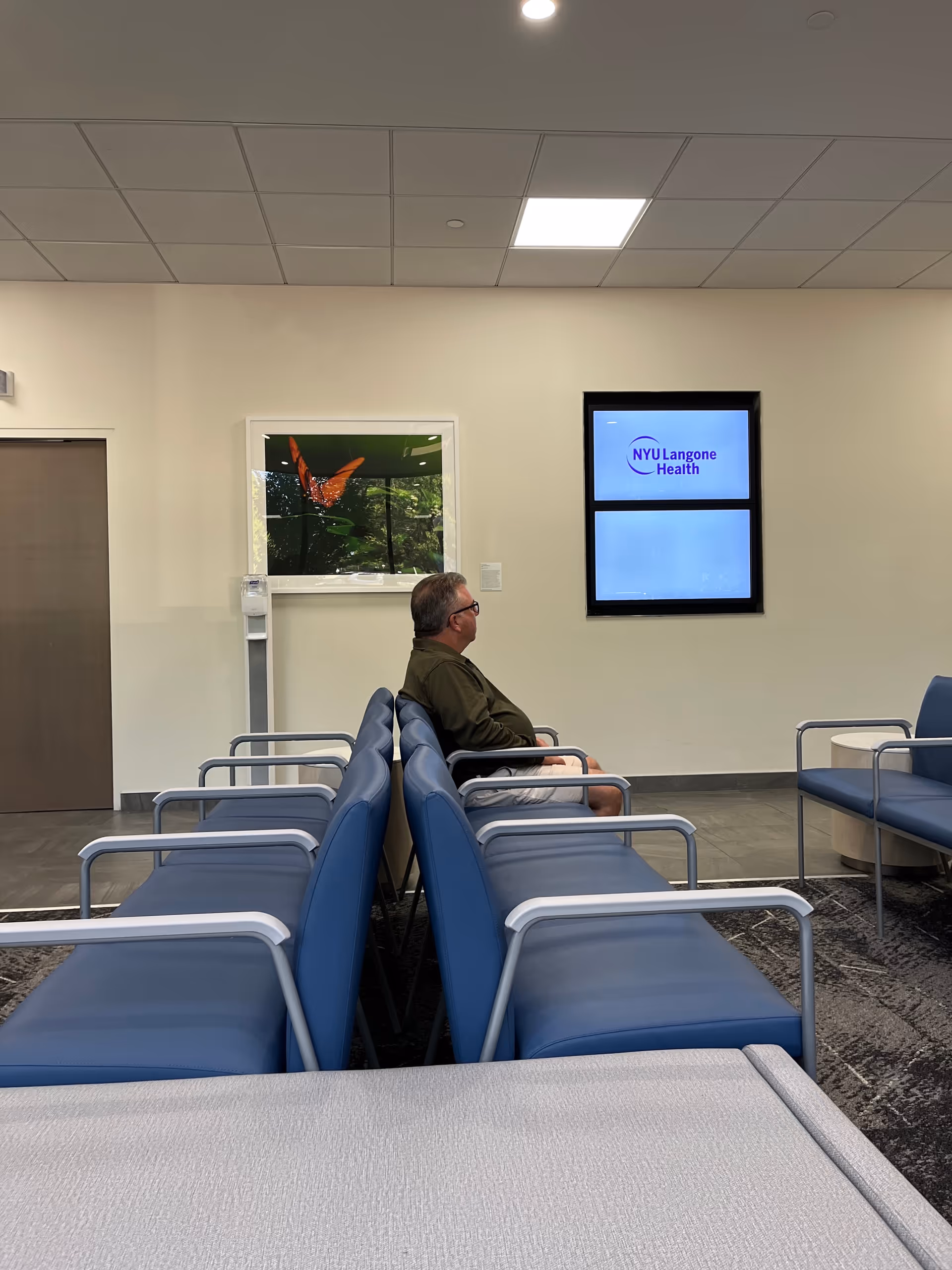Interior waiting area with blue chairs and a man seated, NYU Langone Health sign on wall.