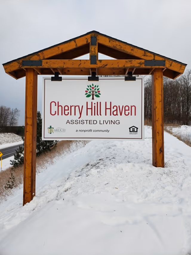 A large wooden sign structure with a peaked roof stands on a snowy hill beside a road. The sign reads 'Cherry Hill Haven ASSISTED LIVING a nonprofit community' with a tree logo above the text and additional small logos at the bottom corners. Leafless trees and a cloudy sky are visible in the background.