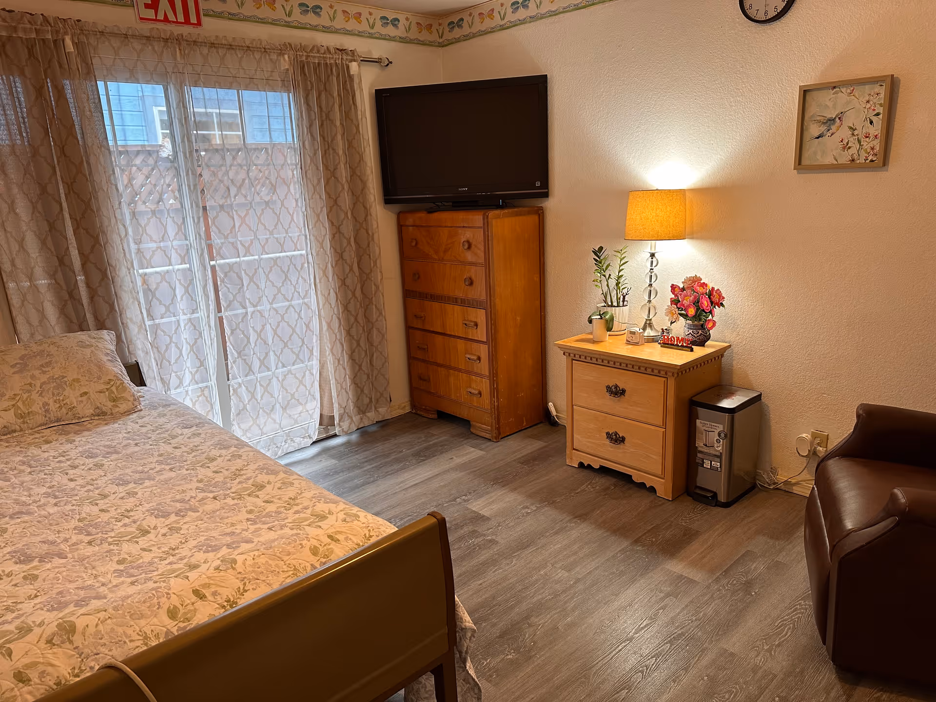 Cozy bedroom with bed, dresser, TV, nightstand, lamp, plants, and sliding glass door.