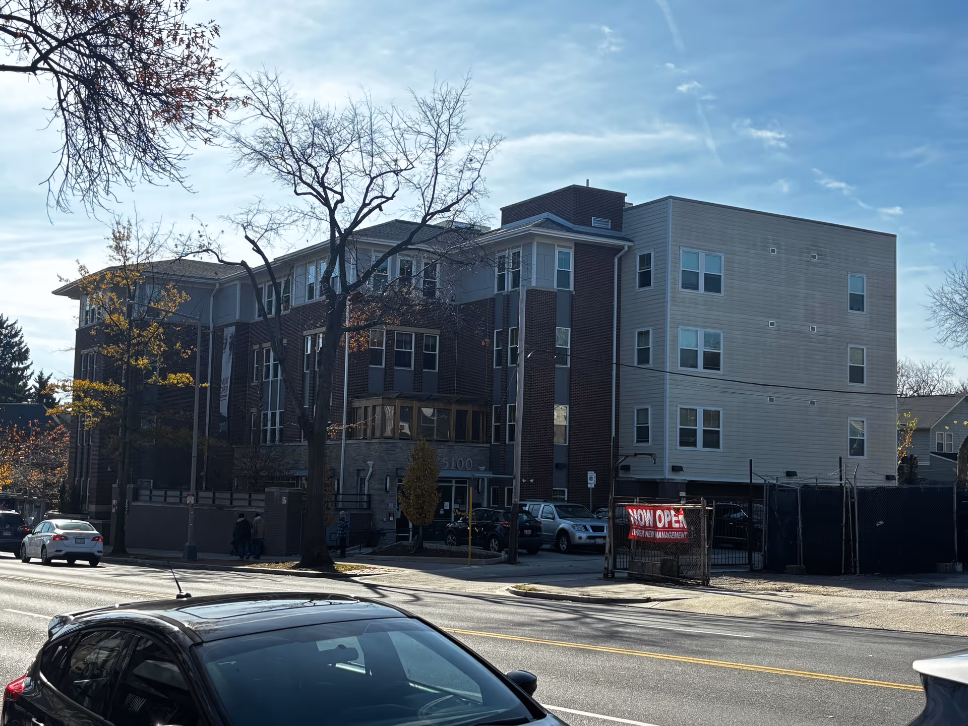 Street view of a three-story senior living building with brick and light siding, leafless trees in front, parked cars, and a 'NOW OPEN' banner.