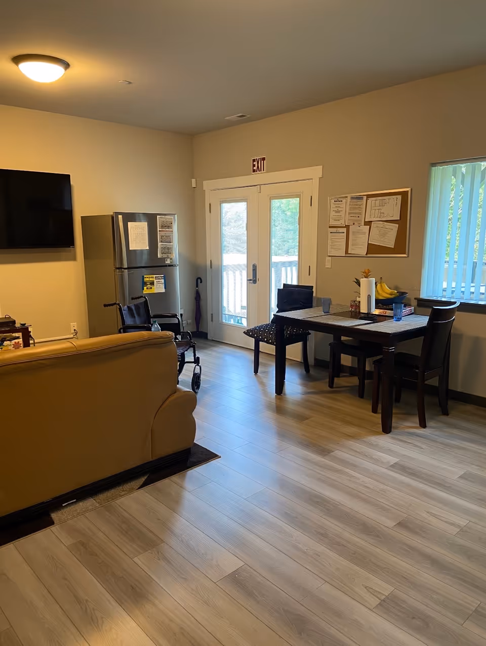 Interior common area with tan sofa, dining table, wheelchair, fridge, TV, and exit door.
