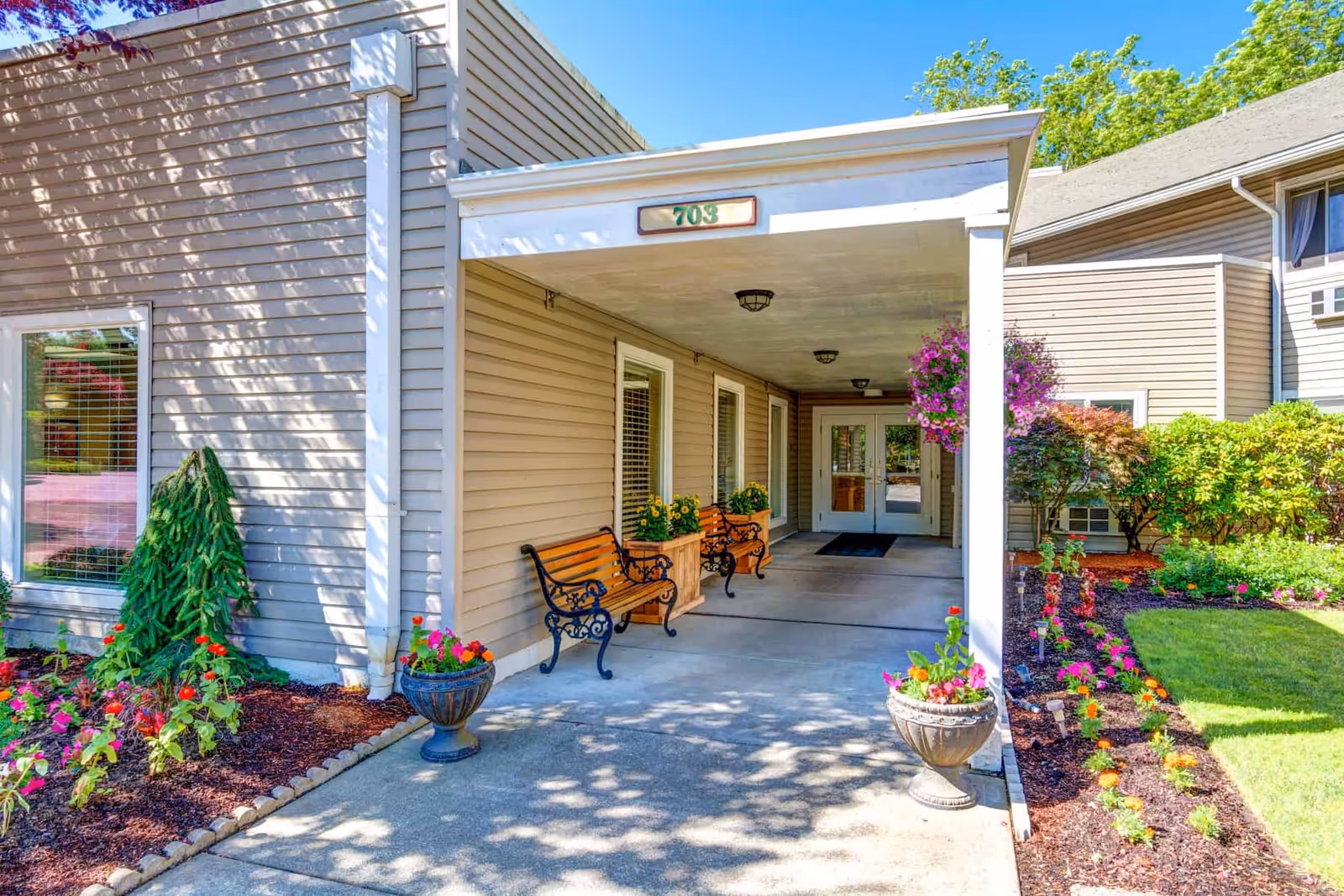 Entrance to a building with beige siding and a covered porch area. There are two wooden benches with black metal armrests and flower planters on either side of the entrance. The porch ceiling has three light fixtures, and the building number 703 is displayed above the entrance. Surrounding the entrance are well-maintained flower beds with colorful flowers and green shrubs under a clear blue sky.