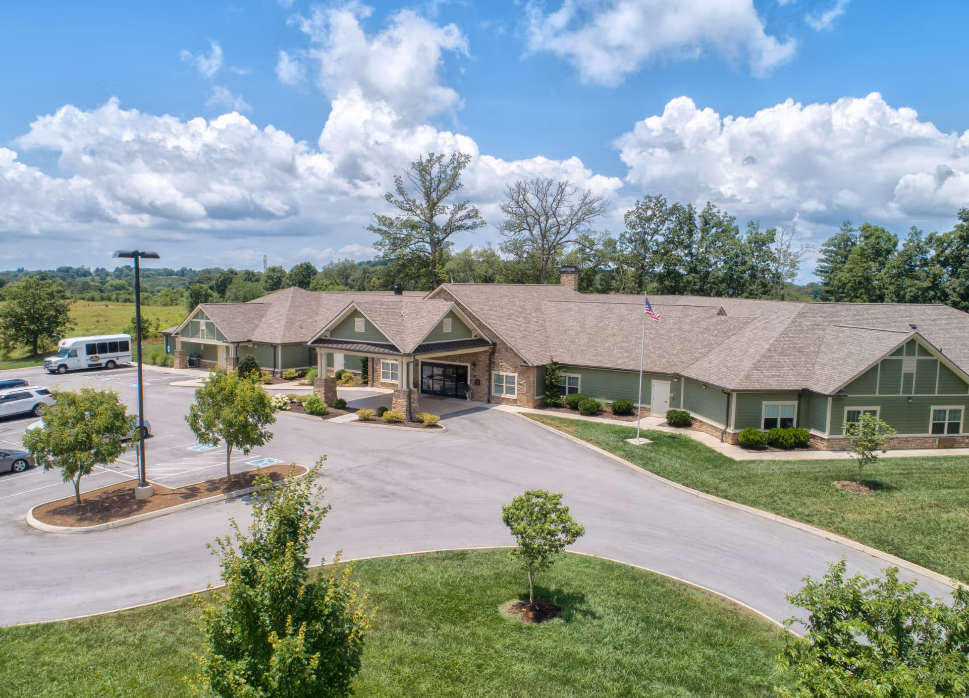 Exterior view of Clover Hill Senior Living facility showing a single-story building with a covered entrance, surrounded by a parking lot with several cars and a shuttle bus, green lawns, trees, and a flagpole with an American flag under a partly cloudy sky.
