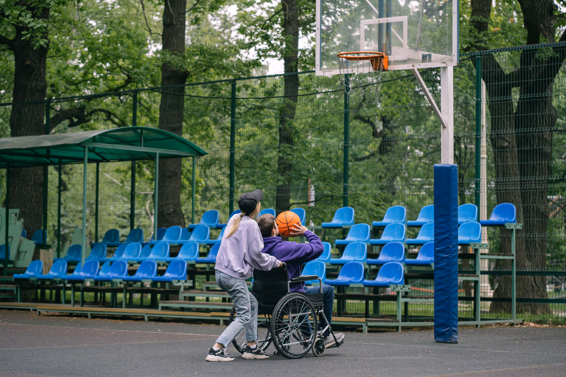 Two people near an outdoor basketball court; one sits in a wheelchair.