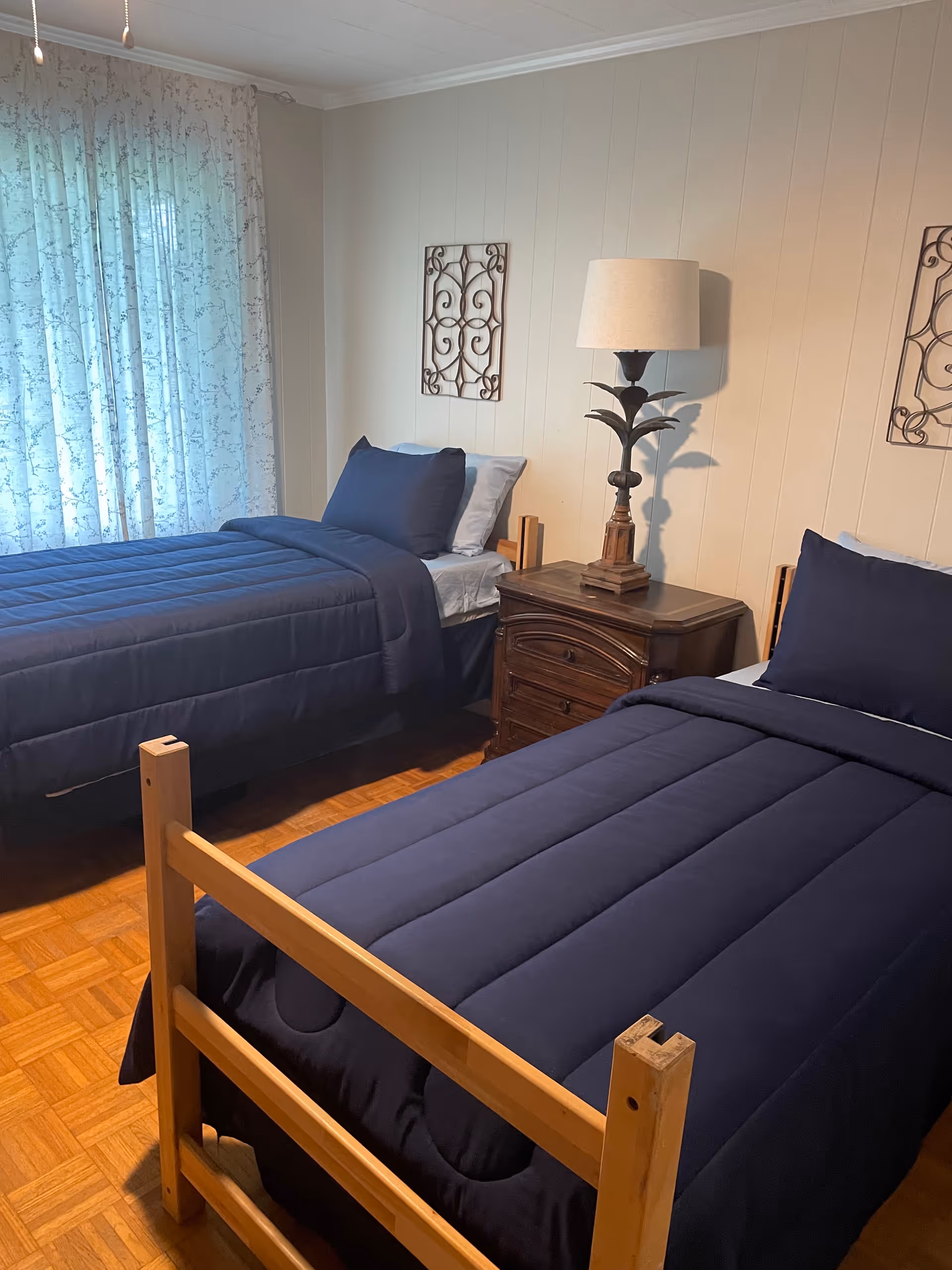 A cozy bedroom with two navy quilted twin beds, a wooden nightstand, and curtains.