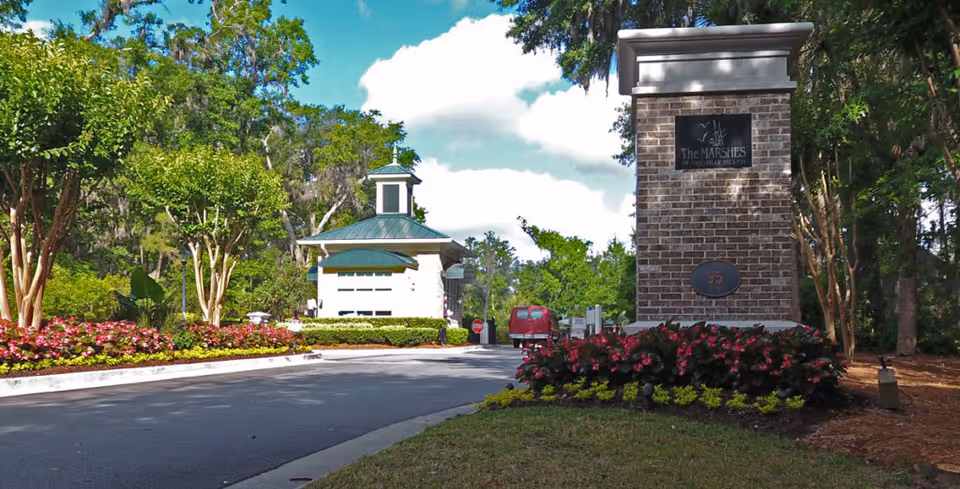 Entrance to The Marshes of Skidaway Island facility with a brick signpost displaying the facility name and number 95, surrounded by landscaped flowers and trees under a partly cloudy sky.