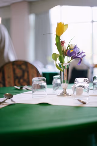 A dining table with a small vase of yellow and purple flowers and upside-down drinking glasses on a white napkin over a green tablecloth.