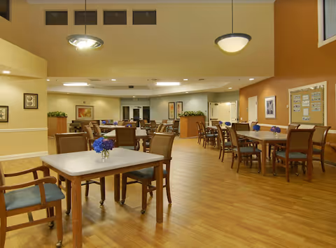 Spacious dining area with multiple wooden tables, chairs, potted plants, and warm overhead lighting.