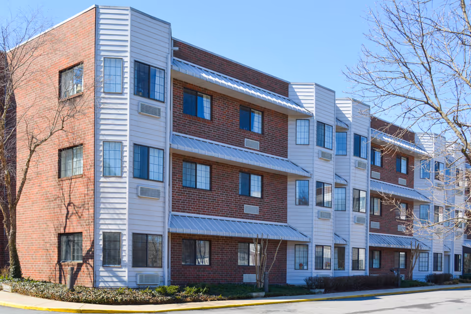 Exterior view of a multi-story senior living facility building with brick and white siding, multiple windows, and small trees in front under a clear blue sky.