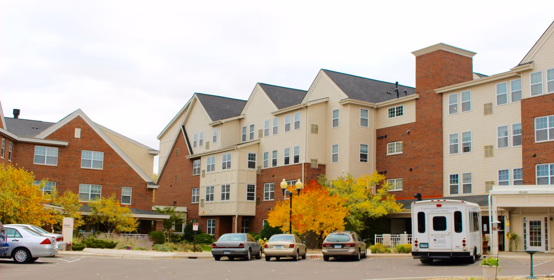 Exterior view of a multi-story senior living facility building with brick and beige siding, several windows, a parking lot with parked cars and a shuttle bus, and autumn-colored trees in front.